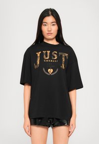 Black oversized t-shirt featuring gold and silver logo print "JUST CAVALLI" with a soft texture, round neckline, and short sleeves.