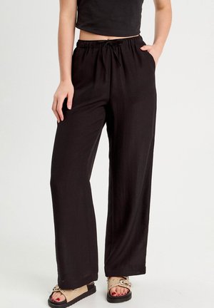 Femme portant un pantalon noir à jambes larges avec taille à cordon de serrage et des sandales beige cloutées, debout devant un fond blanc.