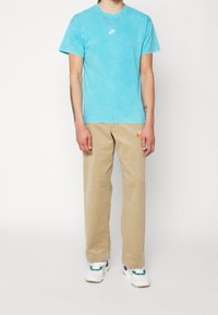 T-shirt en coton turquoise avec le logo Nike, associé à un pantalon en velours côtelé beige arborant un logo Nike orange. Baskets blanches avec des accents verts et bleus.