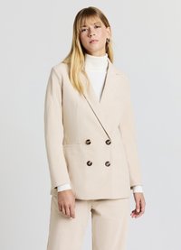 Blazer en velours côtelé beige à double boutonnage avec revers crantés et deux poches avant. Doté de boutons marron foncé et d'une finition lisse et texturée.