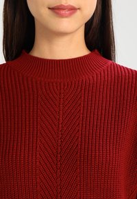 Czerwony sweter robiony na drutach z wysokim prążkowanym dekoltem, teksturowanym wzorem w zygzaki oraz miękkim, przytulnym materiałem. Zbliżenie na dekolt.