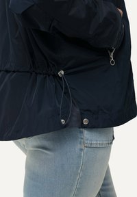 Mörkblå dragkedjajacka i lätt material; elastisk dragsko i midjan och detaljer i silverfärg. Tydlig nätfoder.