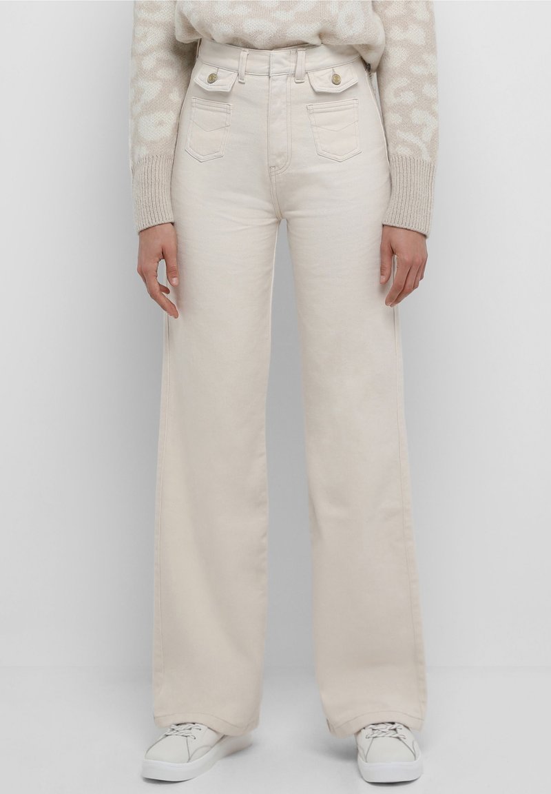 Jean large crème en denim, avec poches avant et accessoires dorés. Porté avec un pull en maille à motifs et des baskets blanches.