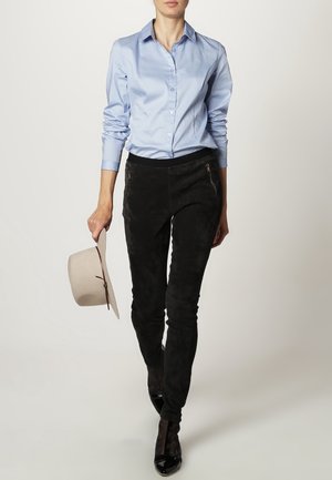 Camisa de botón azul claro combinada con pantalones de ante negros con acentos con cremallera, sostenida por la mano de una persona que lleva tacones negros.