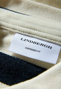 Etykieta Lindbergh z oversized fit na jasnobeżowej tkaninie, teksturowanym dzianinie z ciemnoniebieskimi akcentami. Metka zawiera nazwę marki i szczegóły dotyczące kroju w czarnym tekście.