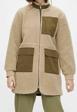 Beige sherpa fleecejakke med olivengrønne stoflommer og brystlomme med lynlås, båret over matchende olivengrønne bukser.