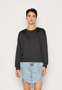 Sweatshirt gris foncé avec des détails en dentelle sur les épaules, col rond et poignets côtelés, associé à une jupe en denim bleu clair avec une fente sur le devant.