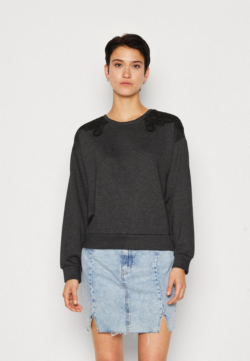 Sweatshirt gris foncé avec des détails en dentelle sur les épaules, col rond et poignets côtelés, associé à une jupe en denim bleu clair avec une fente sur le devant.