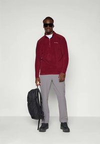 Bordeaux fleece genser med glidelås i kragen, grå bukser, svart ryggsekk og solbriller. Modellen står foran en nøytral bakgrunn.