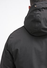 Czarna kurtka z kapturem o gładkiej, matowej fakturze, z przeszyciami i małym logo na tylnej części kołnierza.