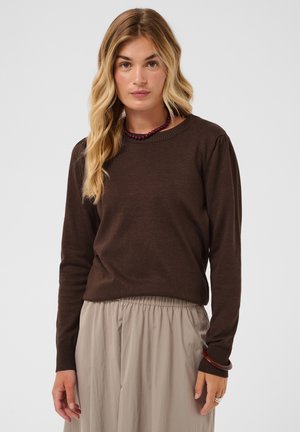 Femme aux longs cheveux blonds portant un pull à manches longues marron foncé et un pantalon ample beige clair sur un fond uni.