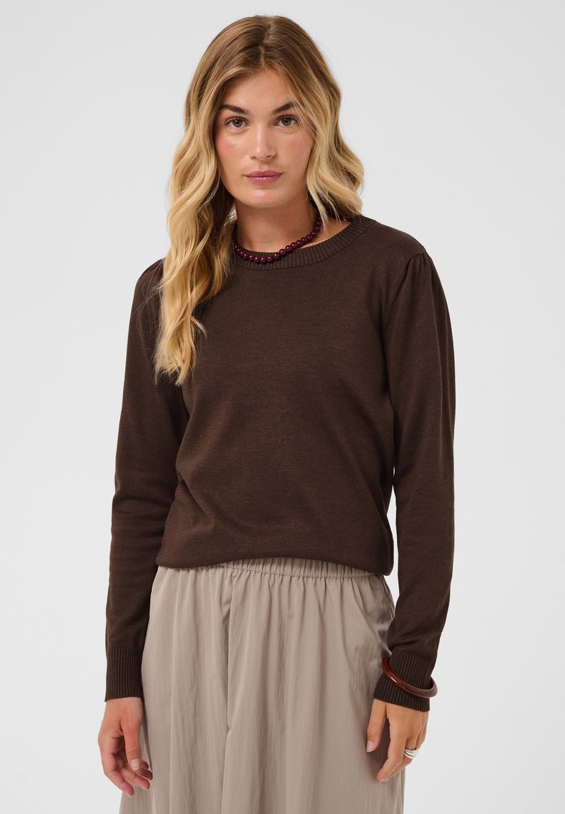 Femme aux longs cheveux blonds portant un pull à manches longues marron foncé et un pantalon ample beige clair sur un fond uni.