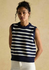 Tumedate ja valged triibulised varrukateta kampsun, millel on soonikkoes kaelus ja alläär, valmistatud pehmest kangast, koos valgete pükstega.