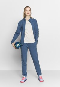 Blauwe sporttracksuit met een ritsjack en broek, uitgerust met patroonzijden, een lichtgrijs T-shirt en meerkleurige sportschoenen.