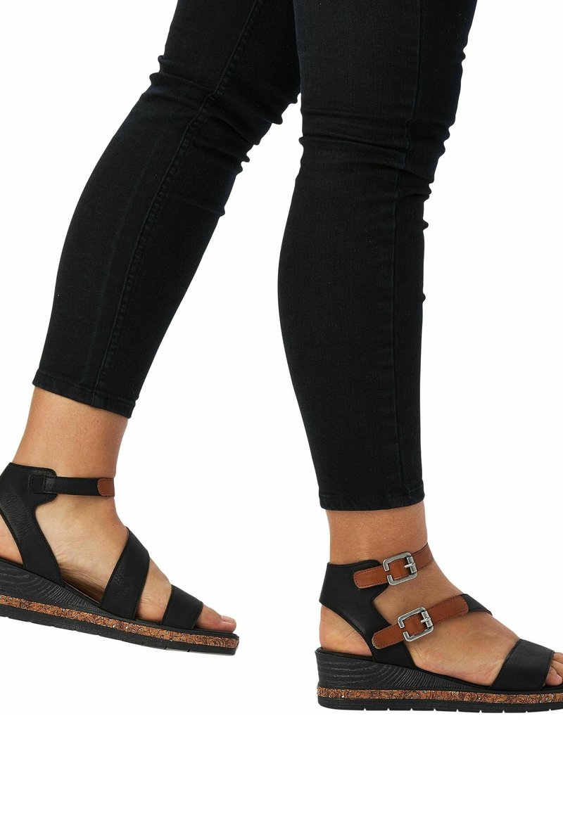 Personne portant un pantalon noir moulant et des sandales compensées noires avec des brides marron et des boucles argentées, un pied levé en plein pas.