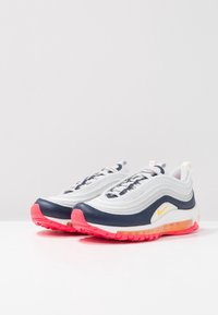 Weißer und marineblauer Sneaker mit strukturiertem Obermaterial, ausgestattet mit einer pinken Gummisohle und Luftdämpfung, reflektierenden Schnürsenkeln und einem gelben Logodetail.