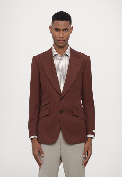 Blazer marron ajusté avec des revers nets, deux poches avant et une fermeture à un bouton, associé à une chemise rayée en dessous.