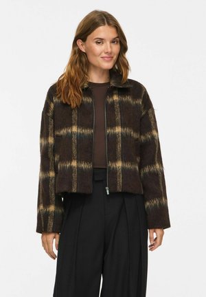 Une femme porte une veste à carreaux marron foncé et beige, zippée sur le devant, avec un pantalon noir à jambes larges, sur un fond clair uni.