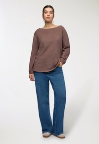 Brązowa, teksturowana bluzka z długim rękawem i dekoltem w łódkę, zestawiona z niebieskimi spodniami o szerokich nogawkach. Brak widocznych wzorów; luźny krój, ciepły materiał.