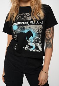 Person wearing a black Linkin Park Meteora band t-shirt with a silhouette graphic, alongside a tattooed arm and black pants.
