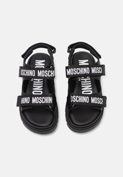 Sandalias negras con doble correa ajustable que presentan el texto blanco "MOSCHINO", suelas de goma, talón abierto y un acabado de textura suave.