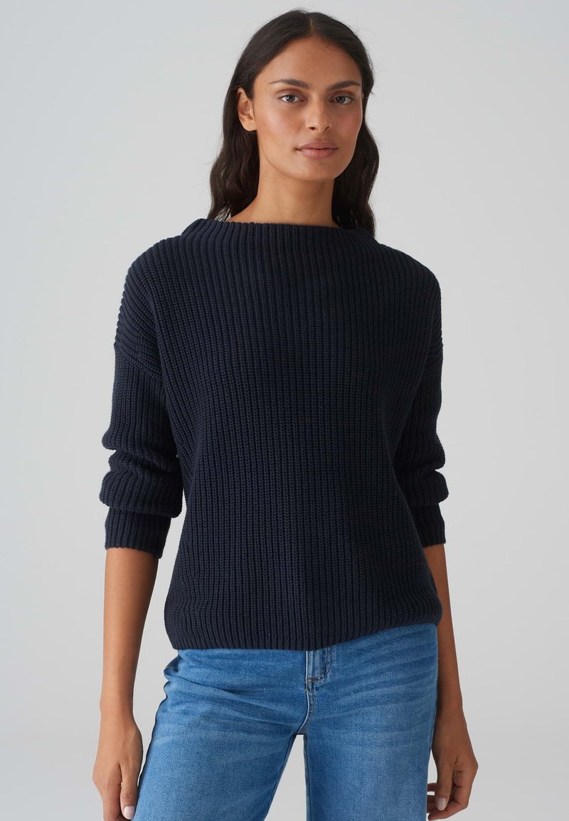 Marineblauer Rippstrickpullover mit weitem Ausschnitt und hochgeschlagenen Ärmeln, kombiniert mit hellblauen Jeans. Strukturierter Stoffdetails sichtbar.