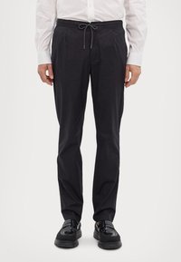 Svarta, lätta byxor med dragsko i midjan, sidofickor och rak passform. Bärs med en vit skjorta med knappar och svarta skor.