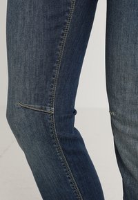 Jeans skinny de denim azul oscuro con un acabado texturizado, que presentan un ajuste slim, costuras sutiles a lo largo del costado y un detalle de bolsillo parcialmente abierto.