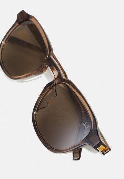 Ochelari de soare cu nuanțe de brun, cu rame geometrice mari, fabricate din plastic transparent. Au accente aurii și lentile închise la culoare.