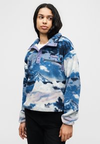 Φλις πουλόβερ με μπλε και λευκό μοτίβο βουνών, μωβ γιακά με κλίπ, τσέπη με φερμουάρ και απαλή υφή, σε συνδυασμό με μαύρο παντελόνι.