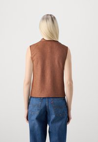 Frau mit glattem, blonden Haar, die ein ärmelloses braunes Top und blaue Jeans trägt und mit dem Rücken zu einer schlichten Wand steht.