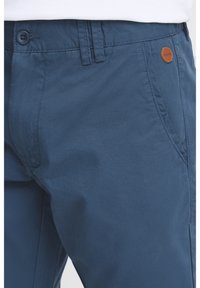 Men's blue cotton blend pants featuring a smooth texture, button closure, and a leather logo accent near the waistline.