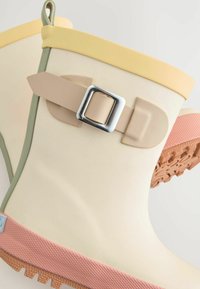 Stivale da pioggia in gomma color crema con bordo giallo e rifiniture verdi. Presenta una cinghia beige con una fibbia argentata e una suola rosa strutturata.