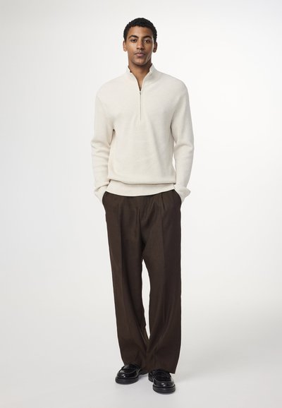 Cremfarbener Pulli mit Reißverschluss, gerippte Textur, kombiniert mit locker sitzenden braunen Hosen und schwarzen Schuhen, vor einem neutralen Hintergrund stehend.