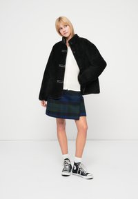 Abrigo de forro polar negro con cierres de palanca, usado sobre un top de punto blanco y una falda de cuadros en azul y verde, combinado con zapatillas altas negras.