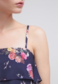 Květinový top s námořnickým pozadím, tenkými popruhy a volánkovými detaily s růžovými, fialovými a zelenými květinovými vzory. Hladká textilie.