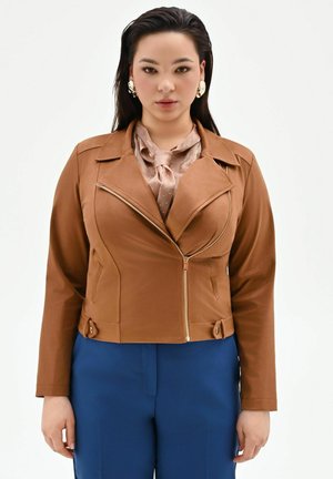 Donna che indossa una giacca marrone con cerniera, camicetta beige a pois annodata al collo, pantaloni blu e grandi orecchini dorati, che guarda in avanti.