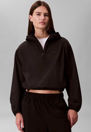 Donna con felpa nera corta con mezza zip e pantaloni con vita elastica neri, in piedi davanti a uno sfondo grigio uniforme.