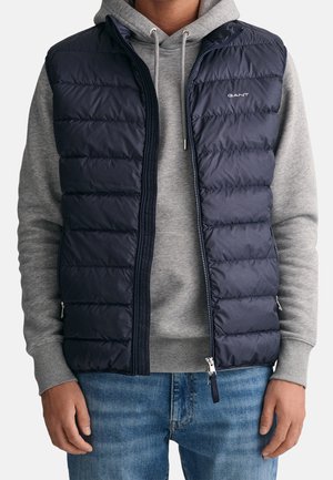 Homme portant une veste matelassée bleu marine par-dessus un sweat à capuche gris et un jean bleu, debout, les bras détendus le long du corps.