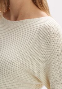 Pull en maille léger de couleur crème, avec une texture côtelée et un col large, fabriqué dans un matériau doux et respirant.
