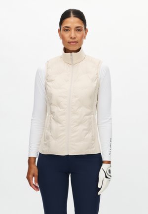 Gilet imbottito beige con collo alto e cerniera frontale. Indossato sopra una maglia bianca a maniche lunghe, abbinato a pantaloni blu navy e guanto da golf.