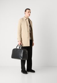 Beige könnyű kabát gombos záródással, fekete bőr táska kettős füllel, fekete nadrág és fekete cipő egy sima háttér előtt.
