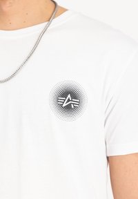 Hvid bomulds T-shirt med et cirkulært sort grafik med et stiliseret logo på brystet, omgivet af et prikket gradient mønster.