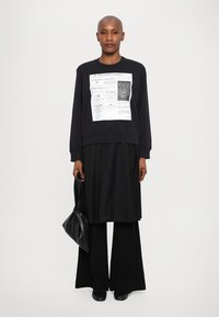 Μαύρη φόρμα με γραφικό τύπωμα μπροστά, φορεμένη πάνω από μια μακριά μαύρη φούστα και φαρδιά παντελόνια, σε συνδυασμό με μια μαύρη τσάντα. Λείος υφασμάτινος. 