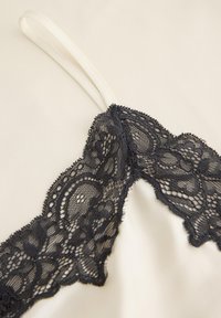Λευκή καμιζόλα με λεπτές τιράντες και μαύρη φλοράλ δαντέλα κατά μήκος του ντεκολτέ και της επάνω περιοχής του στήθους σε ανοιχτό φόντο.