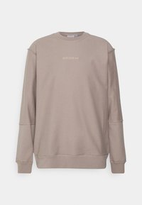 Sudadera marrón clara con cuello redondo, puños acanalados y un pequeño logo bordado en dorado. Textura suave, corte relajado, diseño minimalista.