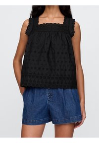 Kvinna som bär en svart ärmlös spetsblus med blommönstrade utskärningar och blå högmidjade denimshorts med framknappar, med händerna i fickorna.