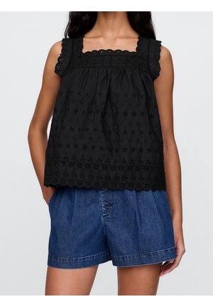 Donna che indossa un top senza maniche nero in pizzo con ritagli floreali e pantaloni corti in denim blu a vita alta con bottoni frontali, mani nelle tasche.