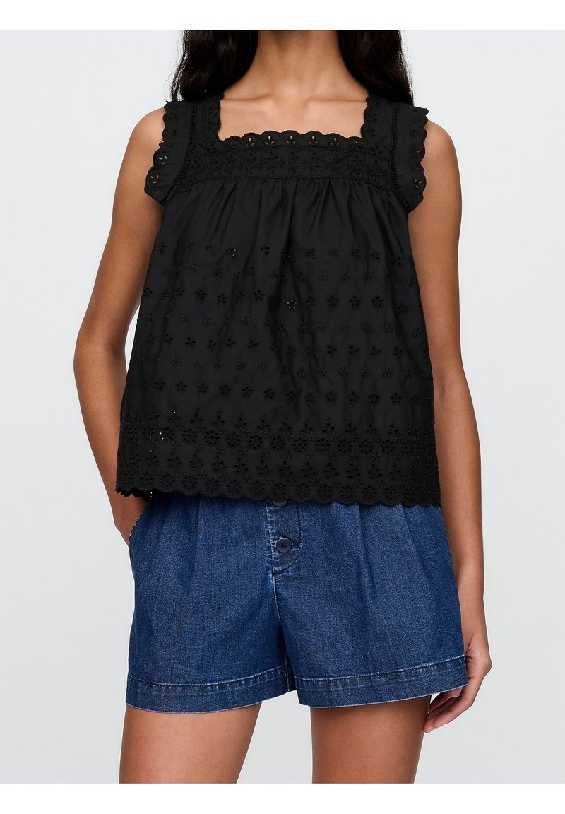 Kvinna som bär en svart ärmlös spetsblus med blommönstrade utskärningar och blå högmidjade denimshorts med framknappar, med händerna i fickorna.