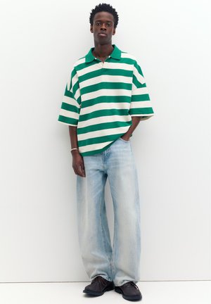 Junger Mann steht vor einer weißen Wand und trägt ein grün-weiß gestreiftes Poloshirt, hellblaue weite Jeans und schwarze Schuhe.